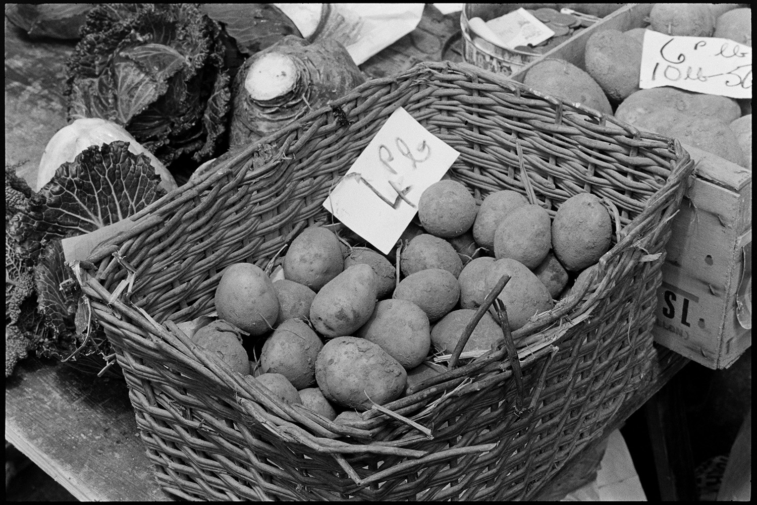 Pannier market, vegetables in baskets, potatoes, swedes, leeks.
[A basket of potatoes, a tray of potatoes, cabbages, a swede, price tickets and a tin with money in it on a vegetable stall at Barnstaple Pannier Market.]