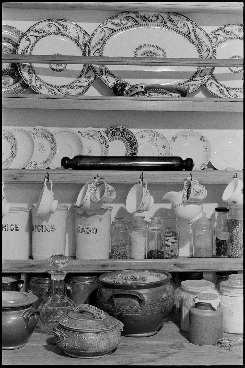 Kitchen utensils, dresser with plates, cups etc.
[Kitchen utensils, plates, storage jars, rolling pin, and cups on hooks displayed on a dresser in Truda Lane's kitchen at Kiverleigh, near Beaford. The image was taken for a cookery book.]