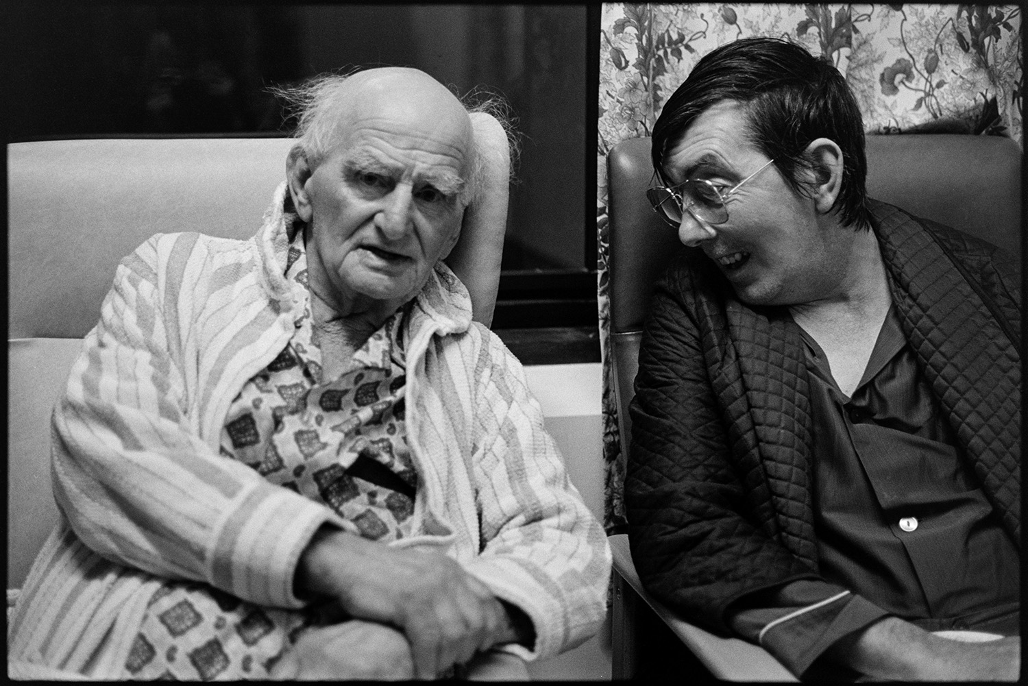 Two elderly men sitting in cottage hospital lounge.
[Archie Parkhouse, on the left, sitting in a chair in dressing gown and pyjamas, and chatting to another man at Torrington Cottage Hospital.]
