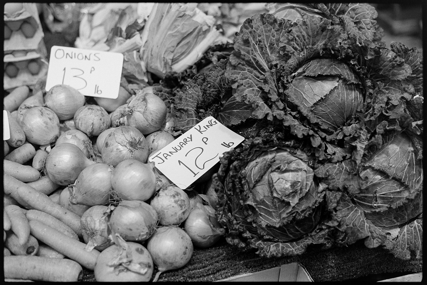 Pannier market, vegetables on display, cabbages, leeks, basket.
[Onions, carrots and January King cabbages on sale at Barnstaple Pannier Market. Price tags are displayed with the vegetables.]