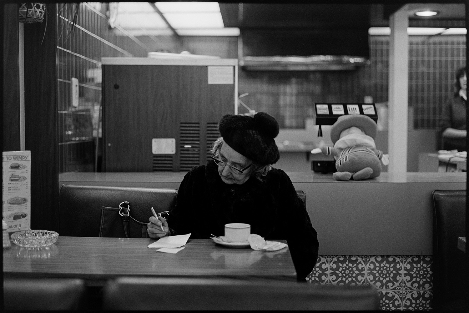 Woman in hat having tea, café.
[A woman wearing a black hat sitting in the Wimpy café in Barnstaple writing a note. An ashtray and cup and saucer is on the table. A toy mascot is on the counter by a drinks machine.]