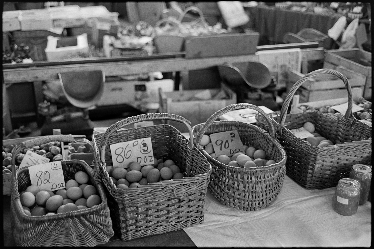 Pannier market, baskets of eggs, vegetables.
[Eggs in wicker baskets for sale at Bideford Pannier Market. Weighing scales, boxes and trays are visible in the background.]