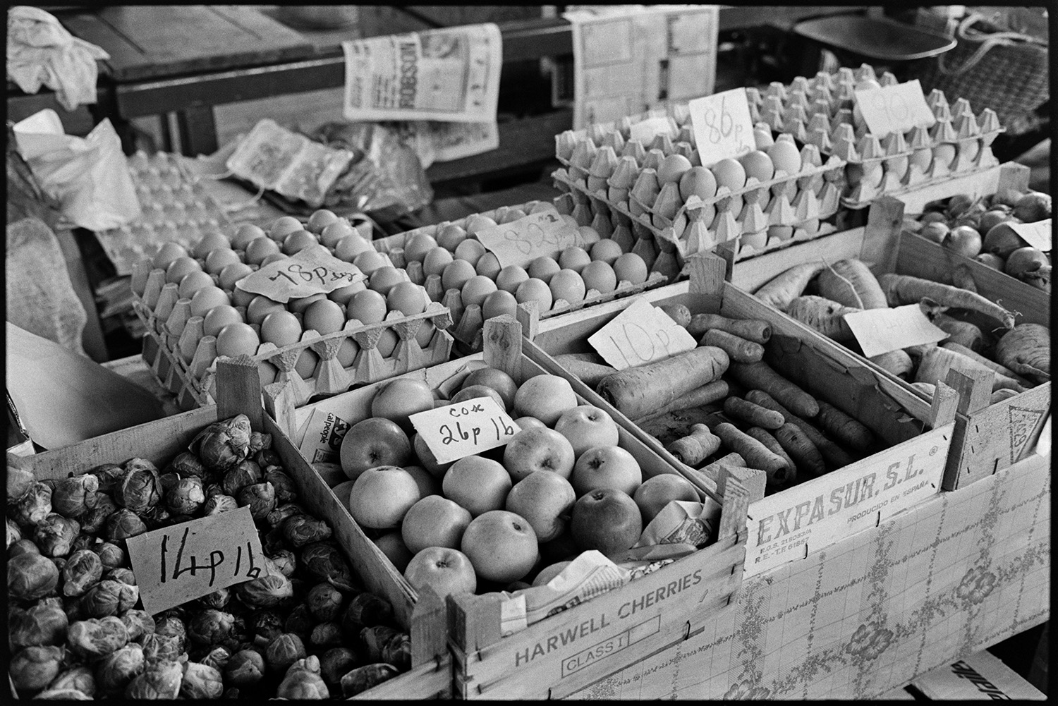 Pannier market, baskets of eggs, vegetables.
[Trays of eggs and boxes of carrots, onions, parsnips, brussel sprouts, and cox apples for sale at Bideford Pannier Market. Price tags are on the produce.]