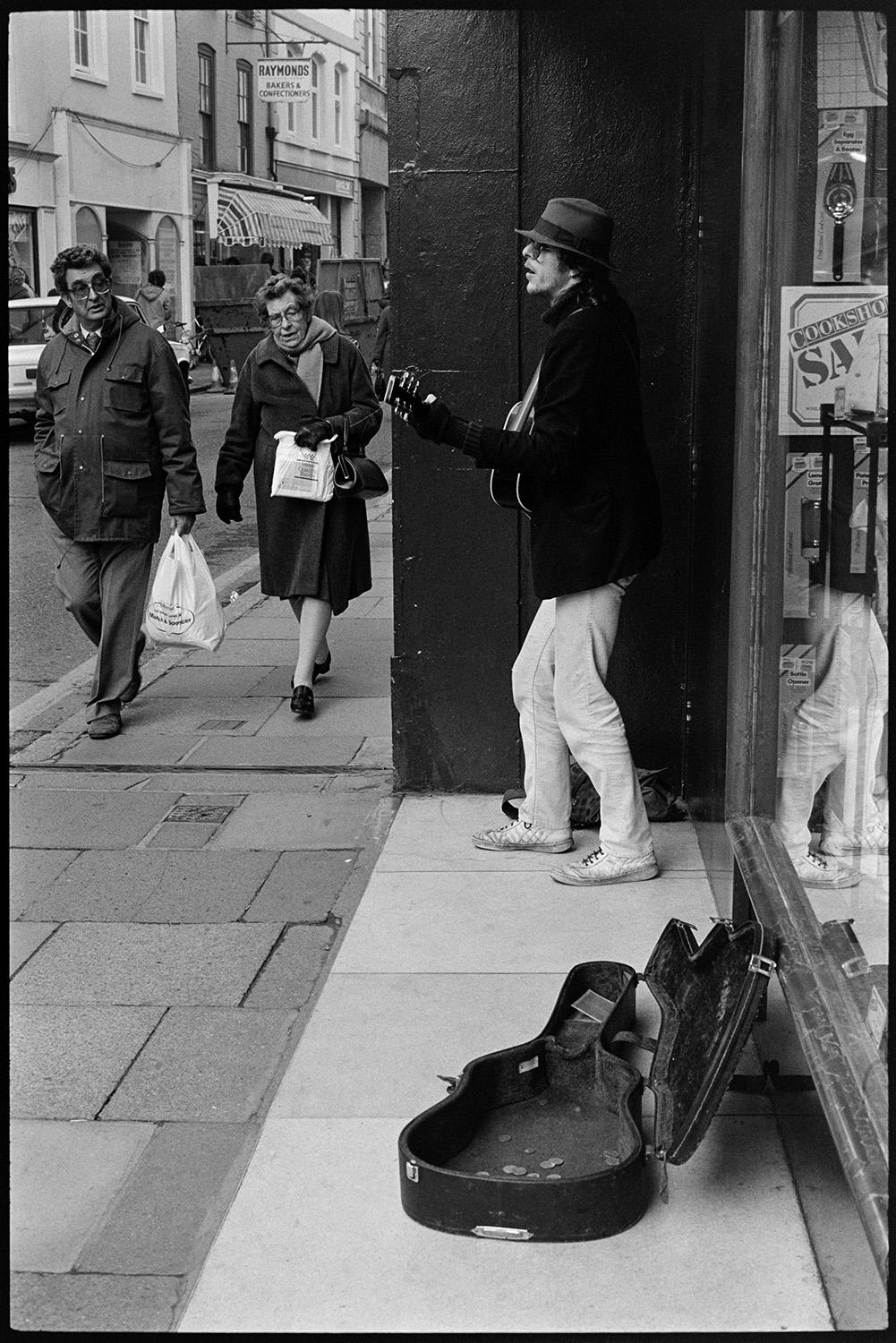 Man busking with a guitar outside a shop on the High Street in Barnstaple. A man and a woman are walking past, carrying shopping. The guitar case is open to collect donations. Raymonds bakery shop is visible in the background.