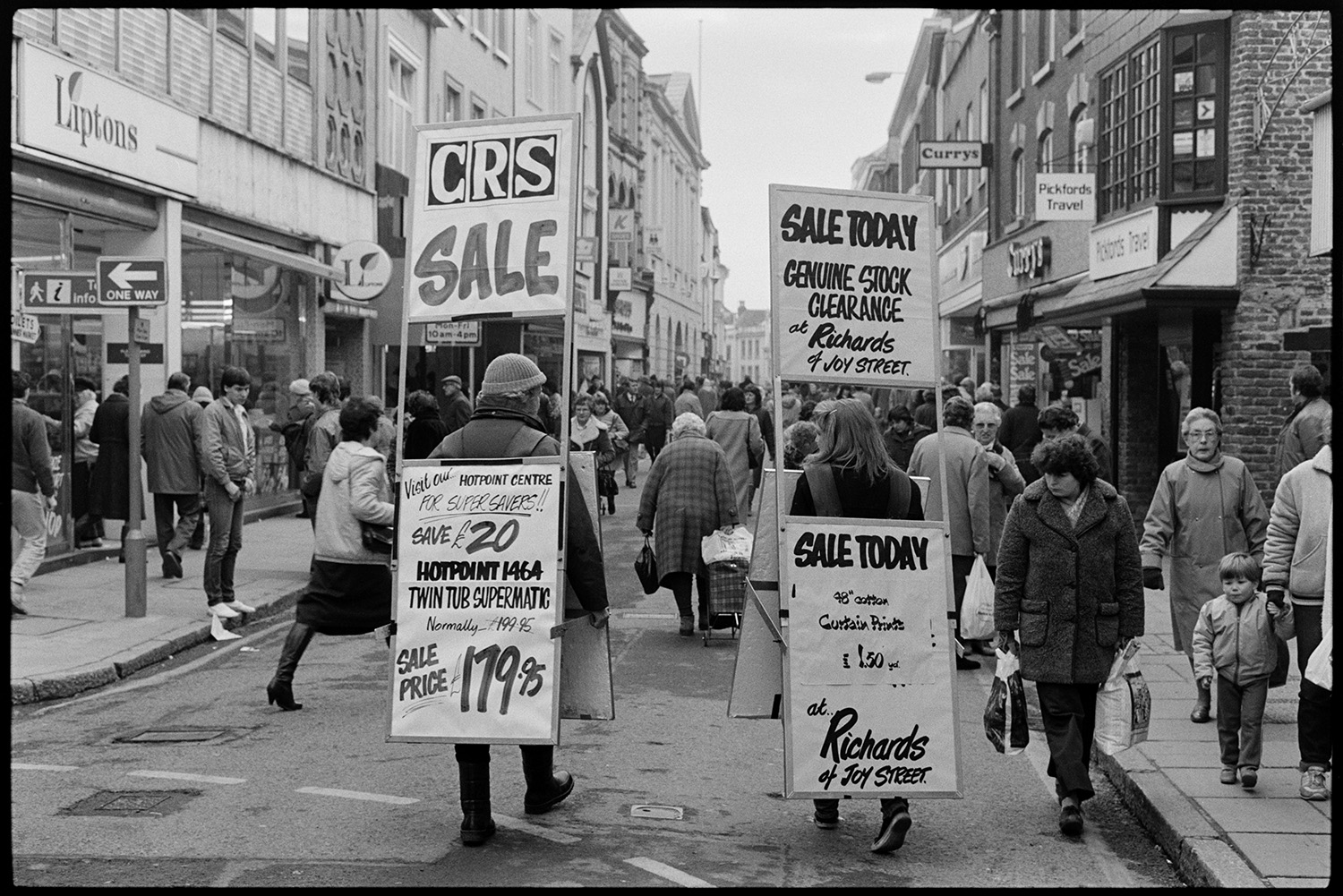 Sandwich persons advertising sales in High street. Sandwich boards.
[Two people walking down the High Street in Barnstaple carrying sandwich boards that advertise sales at Richards and CRS shops. The street is crowded with shoppers, with shops in the background including Liptons, Pickford's Travel and Currys.]