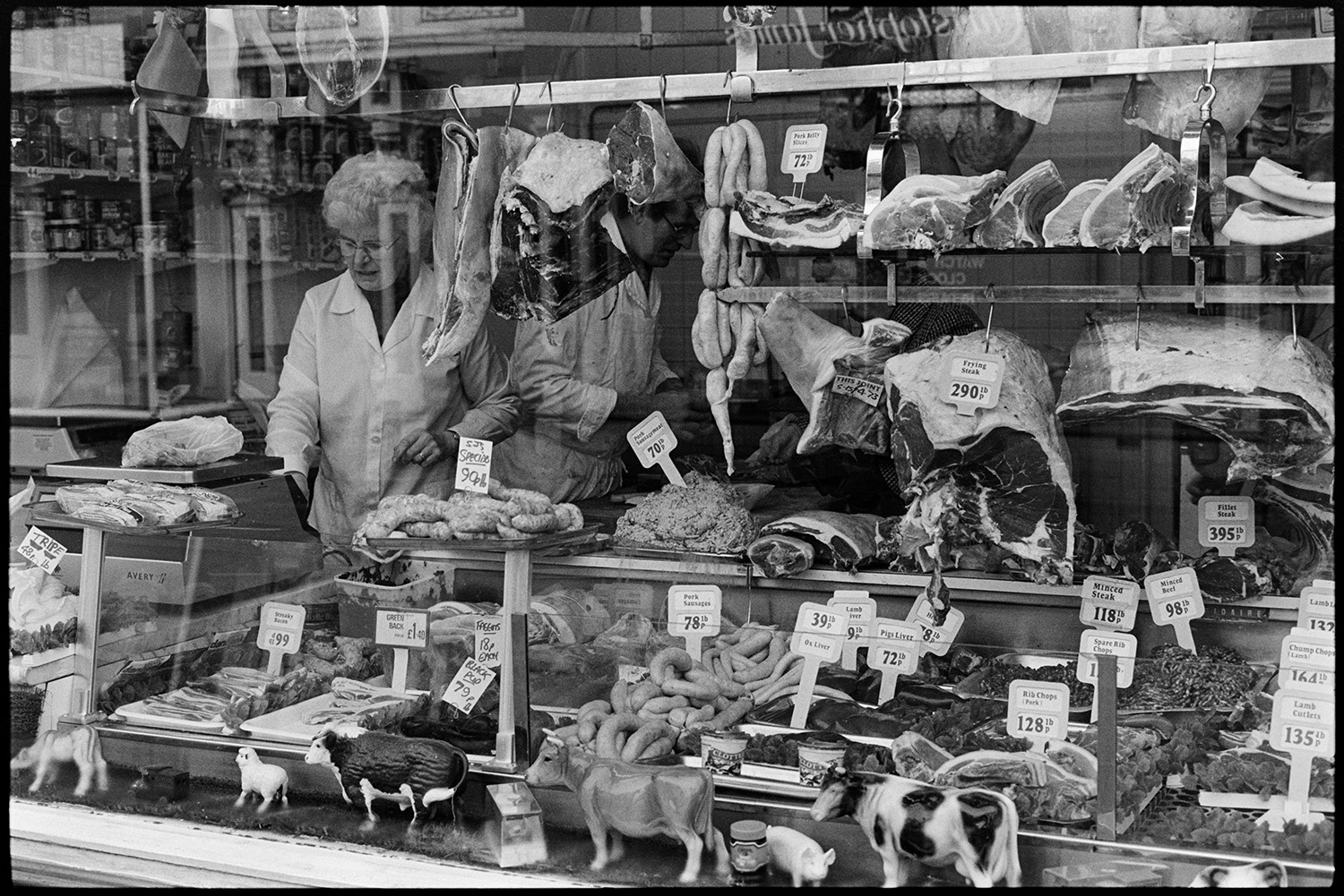 Front of butcher's shop with butcher weighing meat.
[the shop front window of a butcher's shop in Butcher's Row, Barnstaple, with a butcher weighing out meat inside. The shop window is full of meat on display, including sausages, chops, minced beef and steaks. Meat is also shown hanging from hooks with model farm animals on display in the window.]