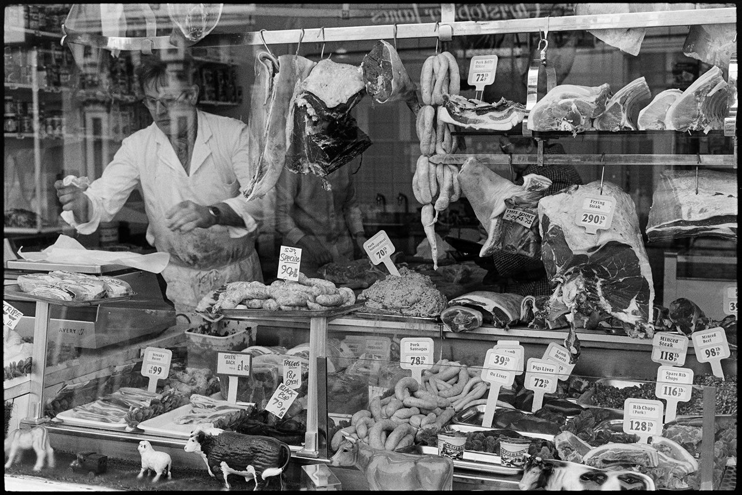 Front of butchers shop with butcher weighing meat.
[The shop front window of a butcher's shop in Butchers Row, Barnstaple, with a butcher weighing out meat inside. The shop window full of meat is on display, including sausages, chops, minced beef and steaks. Meat is also shown hanging from hooks with model farm animals on display in the window.]