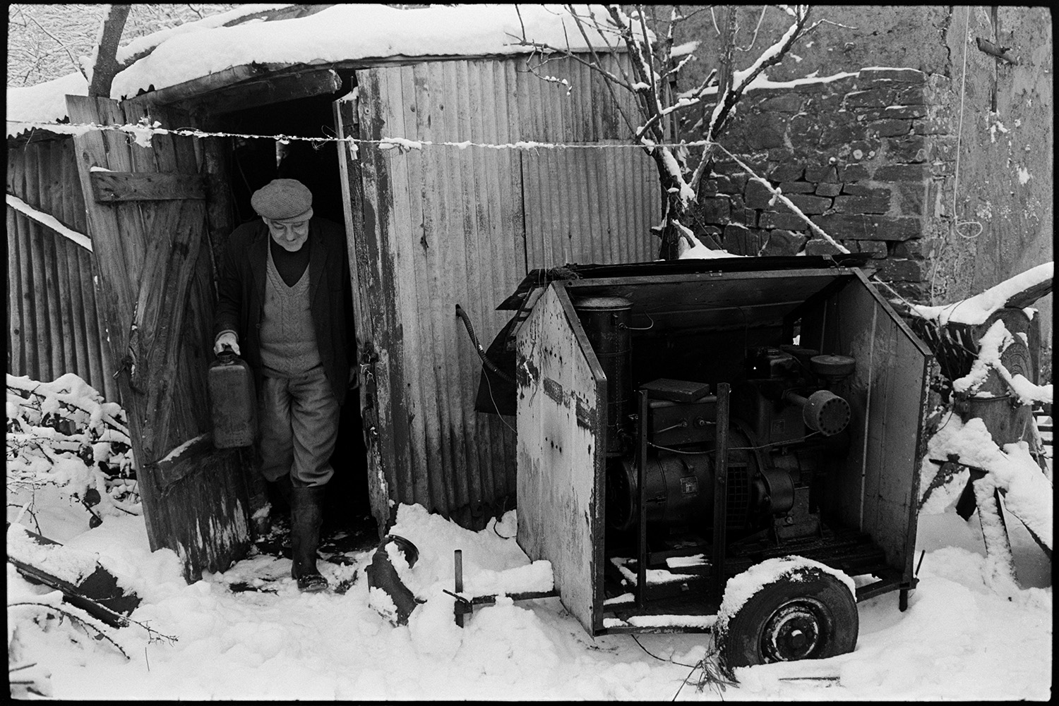 Snow, farmyard poultry, goats and dogs. Tractor and collapsing corrugated iron sheds.
[Cyril Bennett coming out of a corrugated iron shed at Cuppers Piece, Beaford carrying a can of fuel for the generator which is standing outside. The farmyard and shed is covered with snow.]