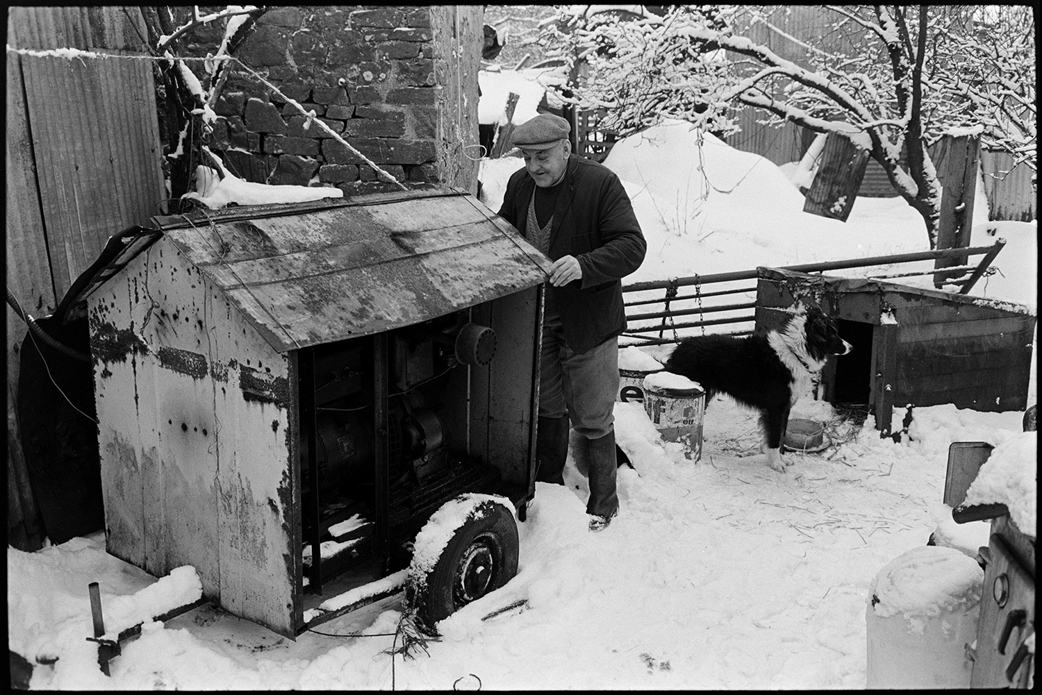 Snow, farmyard poultry, goats and dogs. Tractor and collapsing corrugated iron sheds.
[Cyril Bennett in the snow covered farmyard at Cuppers Piece, Beaford. He is standing by a covered generator which is on wheels, alongside a dog standing by a kennel. There is a metal gate, sheds, and snow covered trees in the background.]