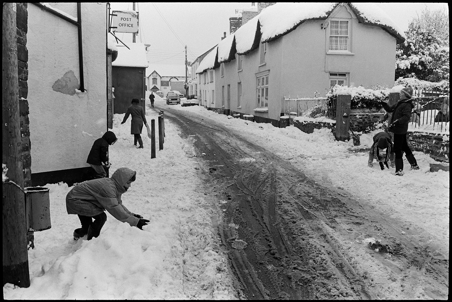 Snow, snowball fight in village street.
[A snowball fight between four children in the Fore Street near the post office in Dolton. The thatched cottage roofs and the road are covered in snow.]