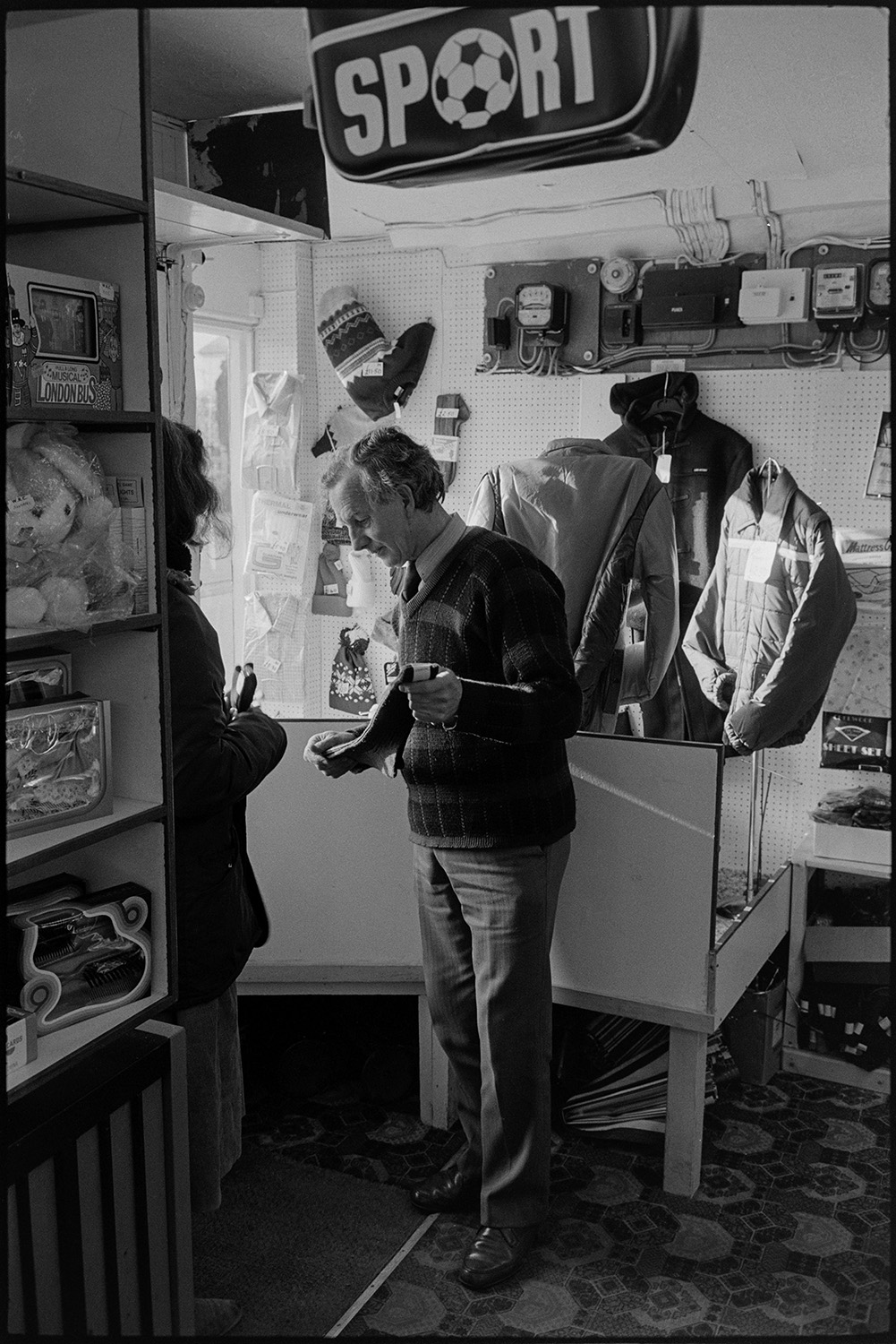 Man selling clothes in shop.<br />
[A man showing a woman a pair of socks at Singh's clothes shop in South Street, Torrington. Shelves with toys and clothes on display, a sports bag, and a counter can be seen inside the shop. An electricity meter is visible on the wall behind the counter.]