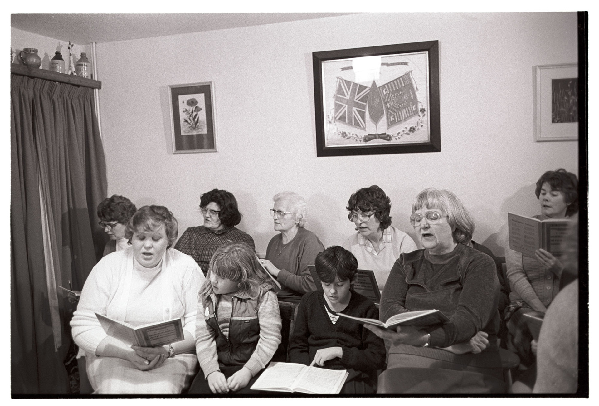 Choir practice in sitting room. Singing. 
[Women and children singing at a choir practice in a living room in a house at St Giles in the Wood. Pictures of a flower and flags are hung on the wall.]