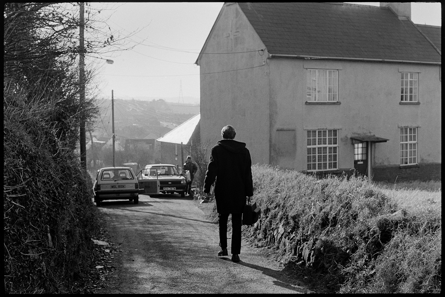 Doctor walking to see patients. 
[Doctor Richard Westcott walking along a road past a house and parked cars, on his rounds to visit patients in South Molton.]