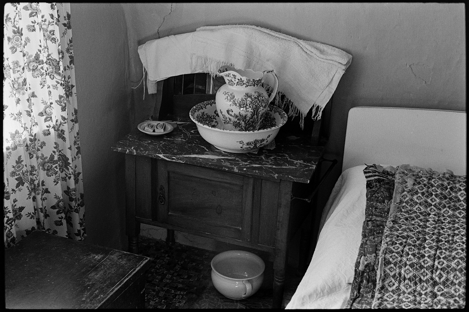 Interior of bedroom, washstand water jug and bowl, chamber pot mirror. Furniture. 
[A wooden washstand with a marble top next to a bed in a bedroom at Bottreaux Mill, Molland. A water jug and bowl, soap an towel are on the stand and a chamber pot is underneath the washstand.]