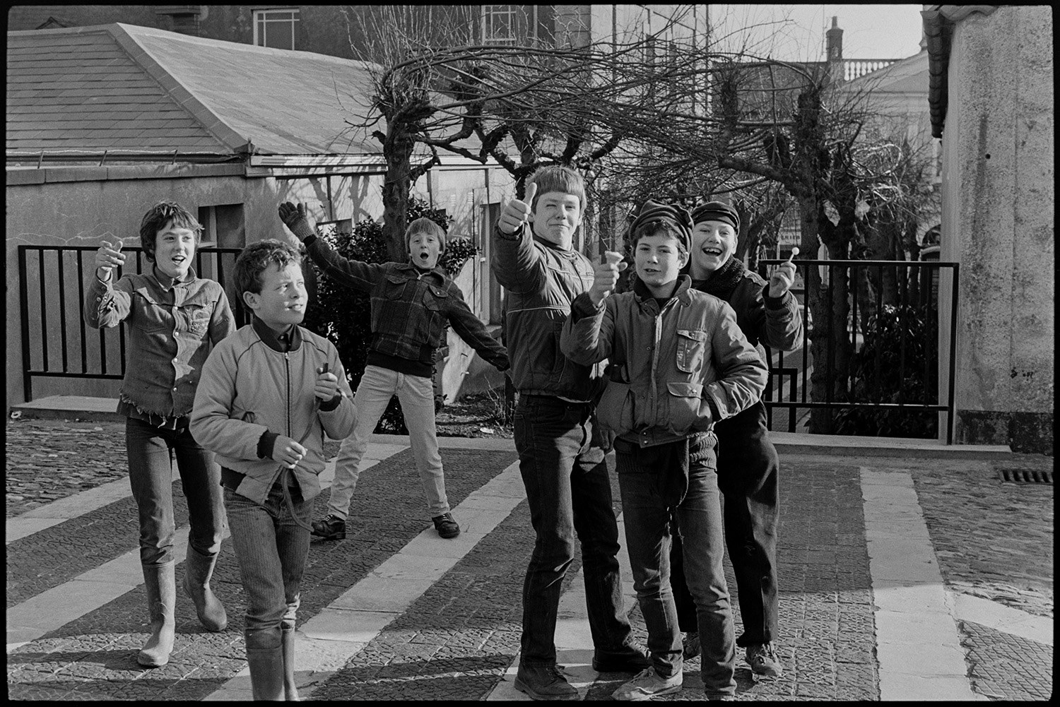 Young boys in churchyard larking in front of camera. 
[A group of six boys waving at the camera on the paved area at the entrance to South Molton churchyard. The avenue of lime trees leading to the churchyard can be seen in the background. Two of the boys are holding an ice cream and a lollipop.]