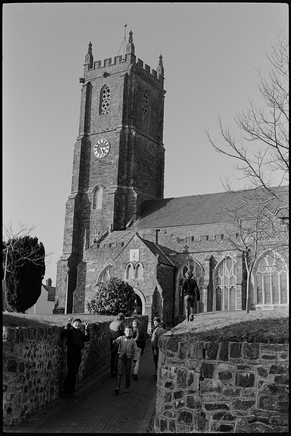 Church with tower. 
[A group of boys walking long the church path through the churchyard towards South Molton Church. Some of them are waving at the camera. The church porch and clock on the church tower are also visible.]