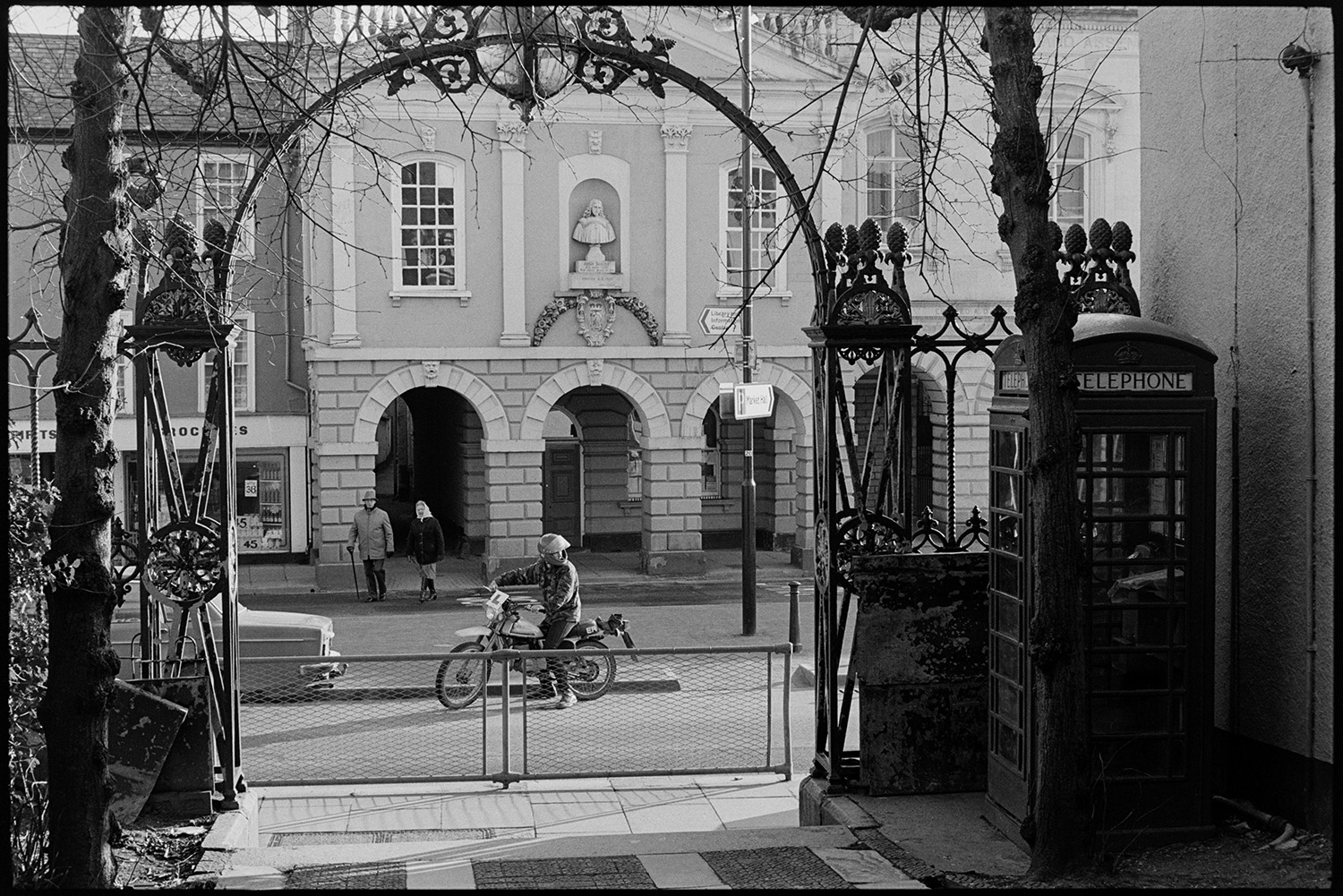 Entrance to avenue of lime trees, wrought iron arch and view of town hall. 
[A wrought iron archway leading to an avenue of lime trees in South Molton. A telephone box can be seen behind the archway. Through the archway South Molton town hall and a motorcyclist about to set off along the street is visible.]