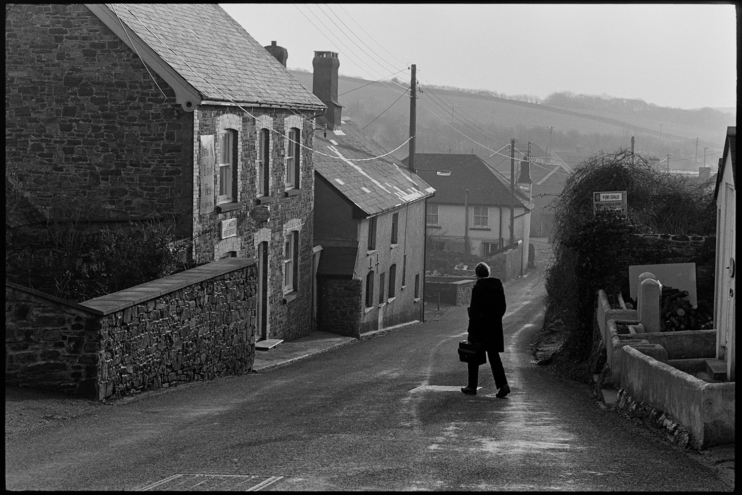 Doctor on his rounds with medical bag. 
[Doctor Richard Westcott walking along a road past houses in North Molton on his rounds to see patients. He is carrying a medical bag.]