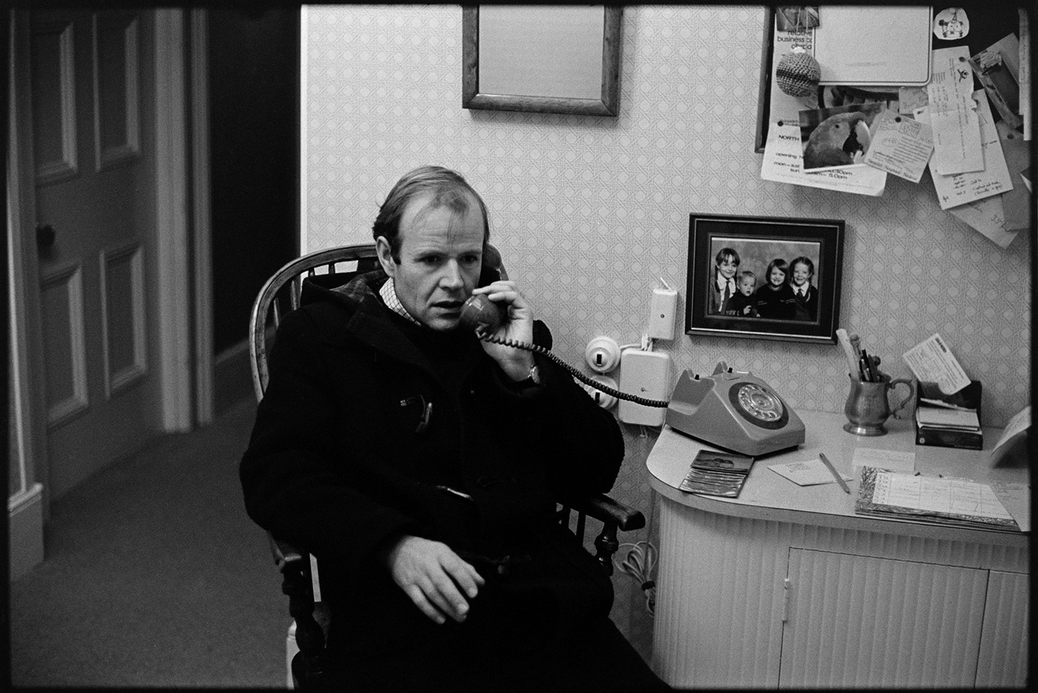 Doctor on his rounds checking man in bed with emphysema. Also telephoning. 
[Doctor Richard Westcott making a call on a telephone possibly in a patients house at North Molton. Various papers are pinned to a notice board above  family photograph on the wall in the background.]