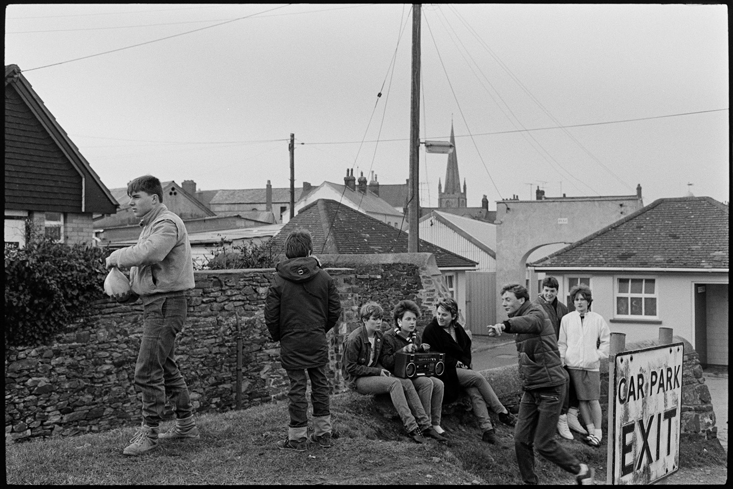 Teenagers sat on a grassy bank by a wall in a car park. One of them is holding a stereo and another is walking across the bank holding a water balloon. Rooftops can be seen in the background.