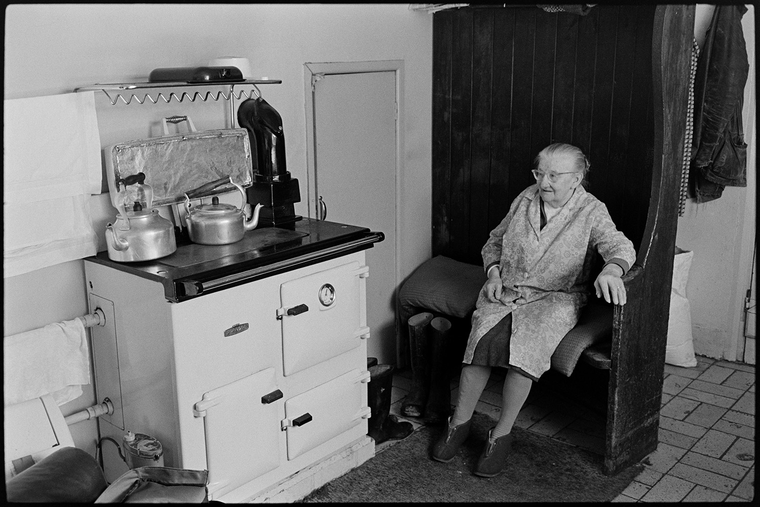 Elderly woman sitting beside Rayburn stove in kitchen. Kettles, china dishes. 
[A woman sitting on a settle bench by a rayburn stove in a kitchen. Two kettles are on the stove.]