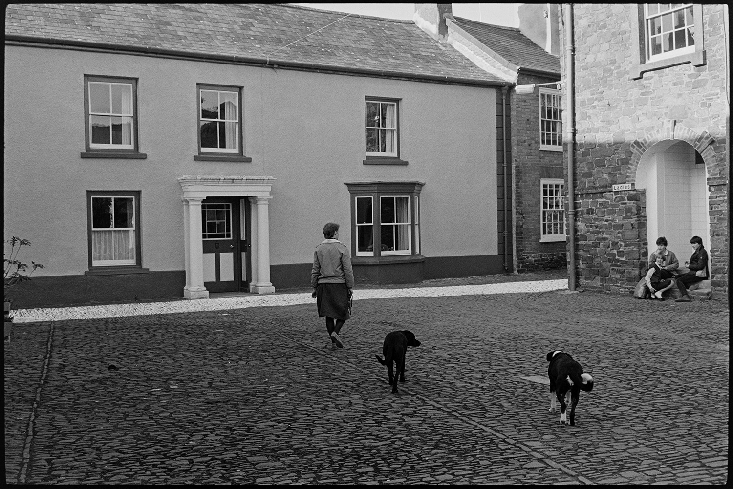 House and cobbled square, former school, porch. 
[A woman and two dogs walking along a cobbled square in front of a house, formerly the School House, in Chulmleigh. The front door has an entrance porch with two pillars. Three young girls are sat outside the entrance to the ladies toilets in the square.]