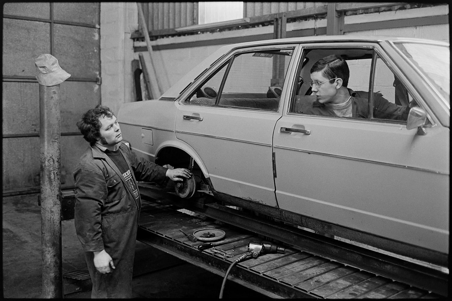 Men working in garage, tools, oil cans, tyres, mending puncture. 
[Ron Turner changing a tyre on a car on a ramp at Dolton Beacon Garage.  He is talking to a person sat in the drivers seat of the car.]