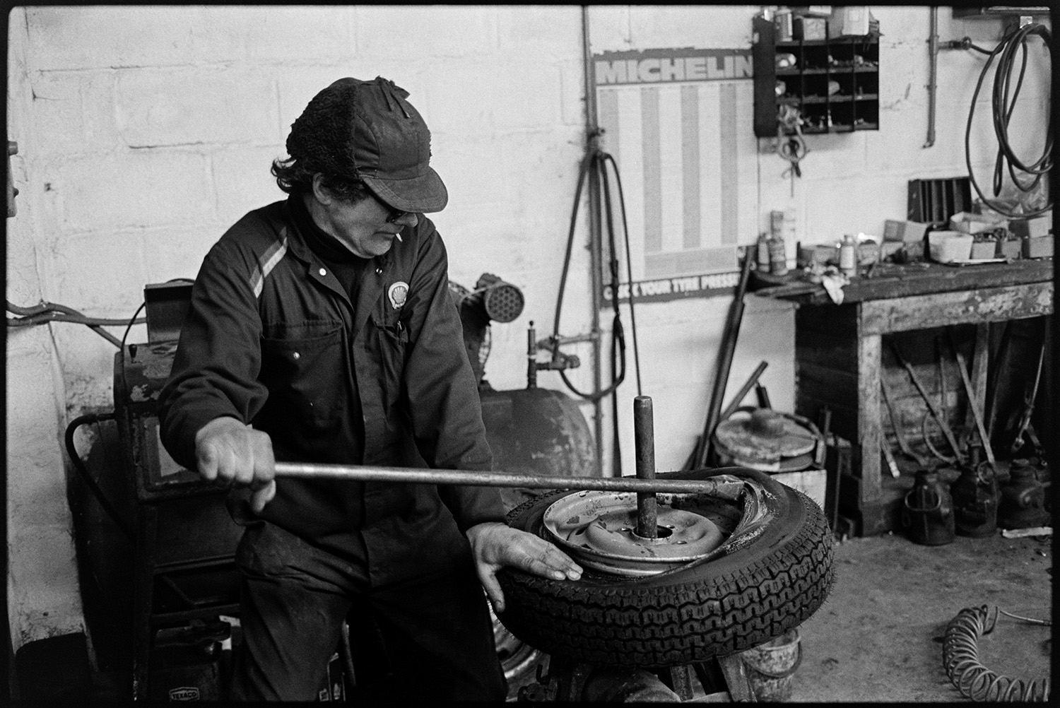 Men working in garage, tools, oil cans, tyres, mending puncture. 
[A man putting a new tyre on a wheel at Dolton Beacon Garage.]