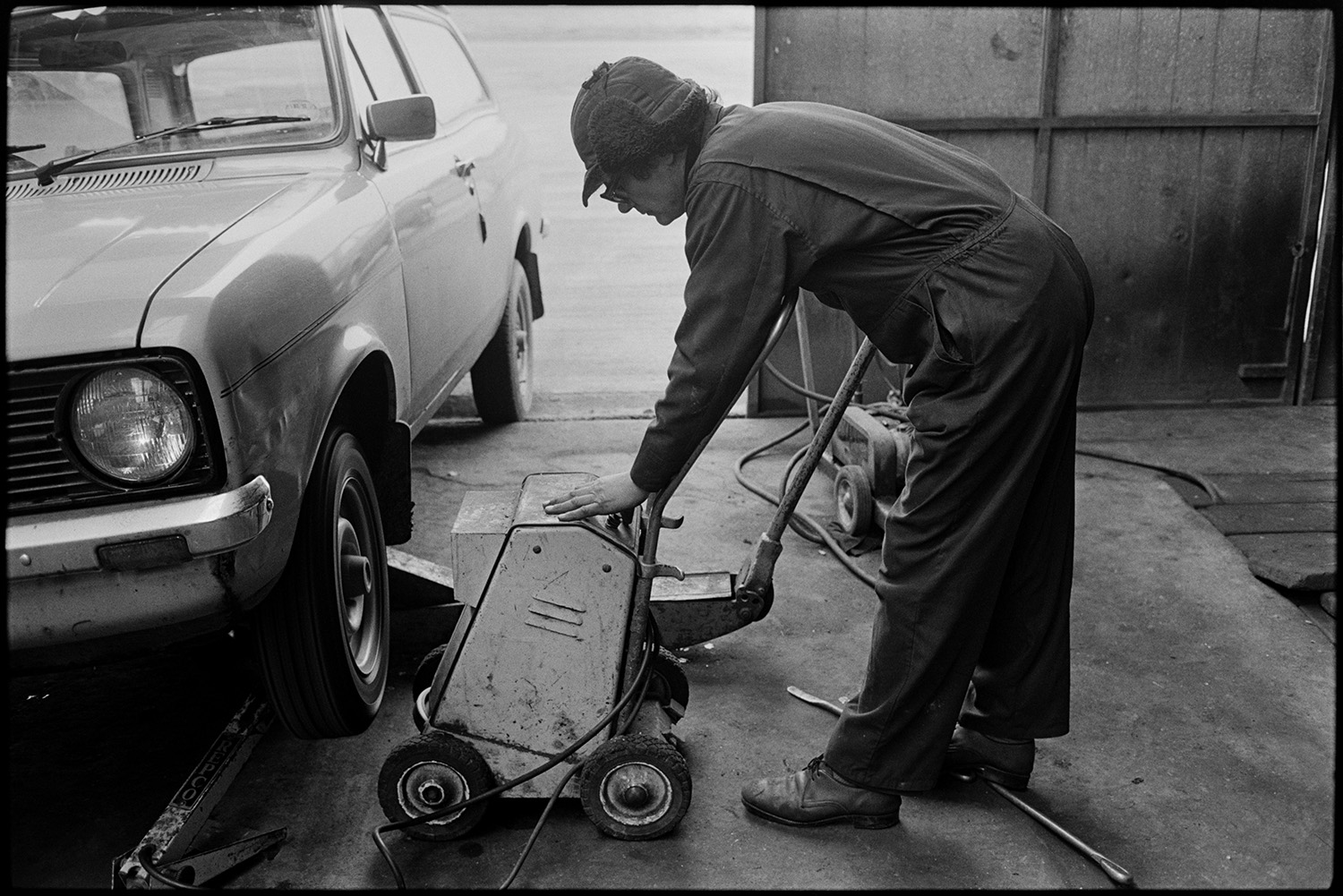 Men working in garage, tools, oil cans, tyres, mending puncture. 
[A man putting a wheel and new tyre back onto a car after fixing a puncture at Dolton Beacon Garage.]