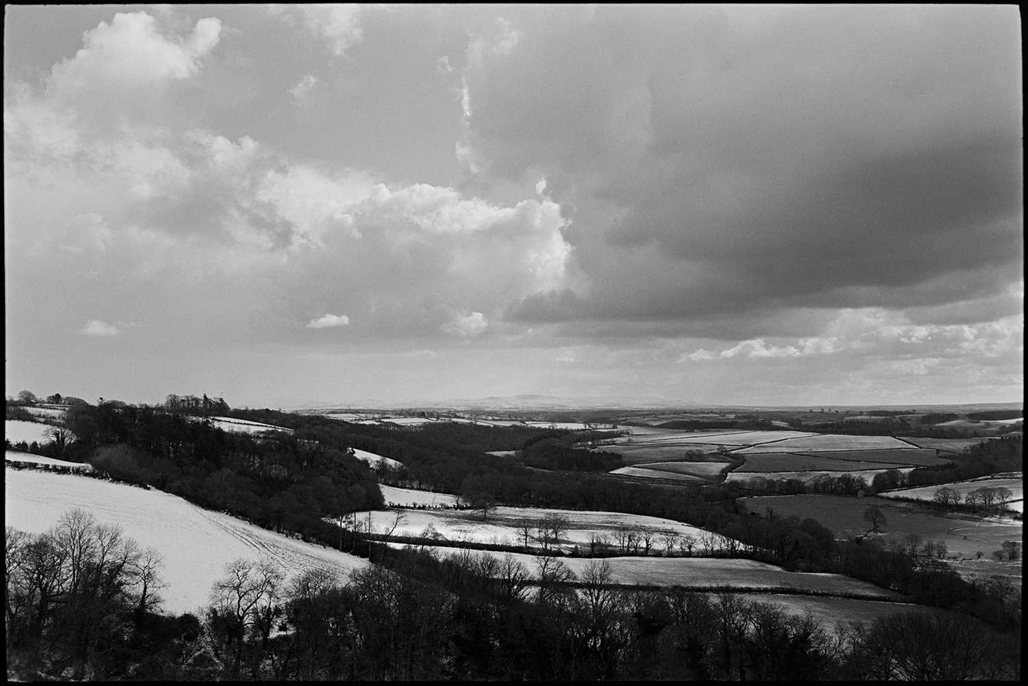Snow, cloudy landscape with distant moor. 
[A landscape with snow covered fields, trees and hedgerows at Harepath, Beaford. Clouds are visible in the sky above.]