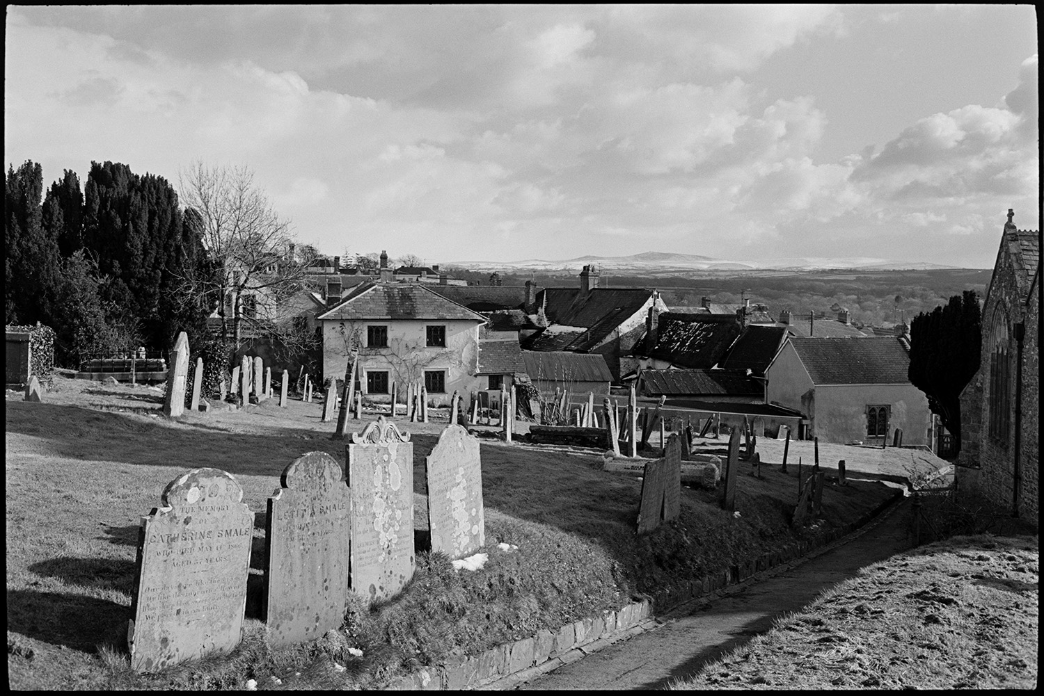 View across graveyard with houses and distant moor. 
[A view of Hatherleigh churchyard with gravestones and the church path. Houses and rooftops can be seen in the background and Dartmoor is visible on the horizon.]