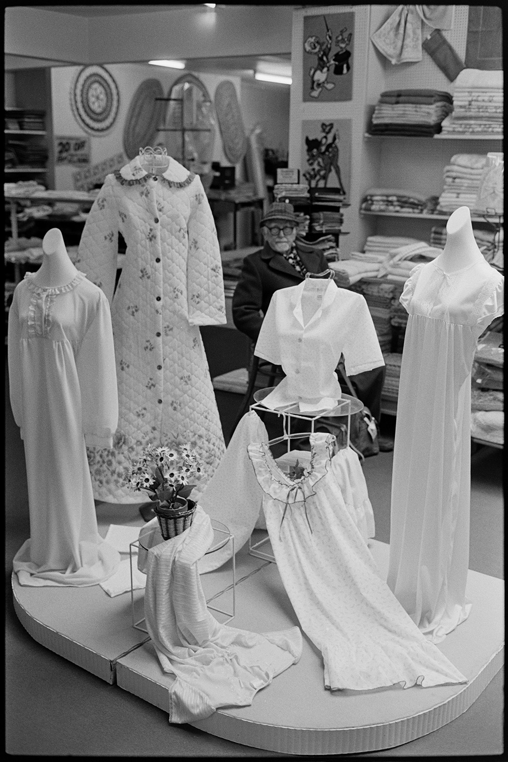 Clothes display in shop, ladies, women's nightwear, dressing gown. 
[A display of women's nightwear, including a dressing gown, in Trapnells clothes shop in Bideford High Street. A man sat on a chair and more clothes stacked on shelves can be seen in the background of the shop.]