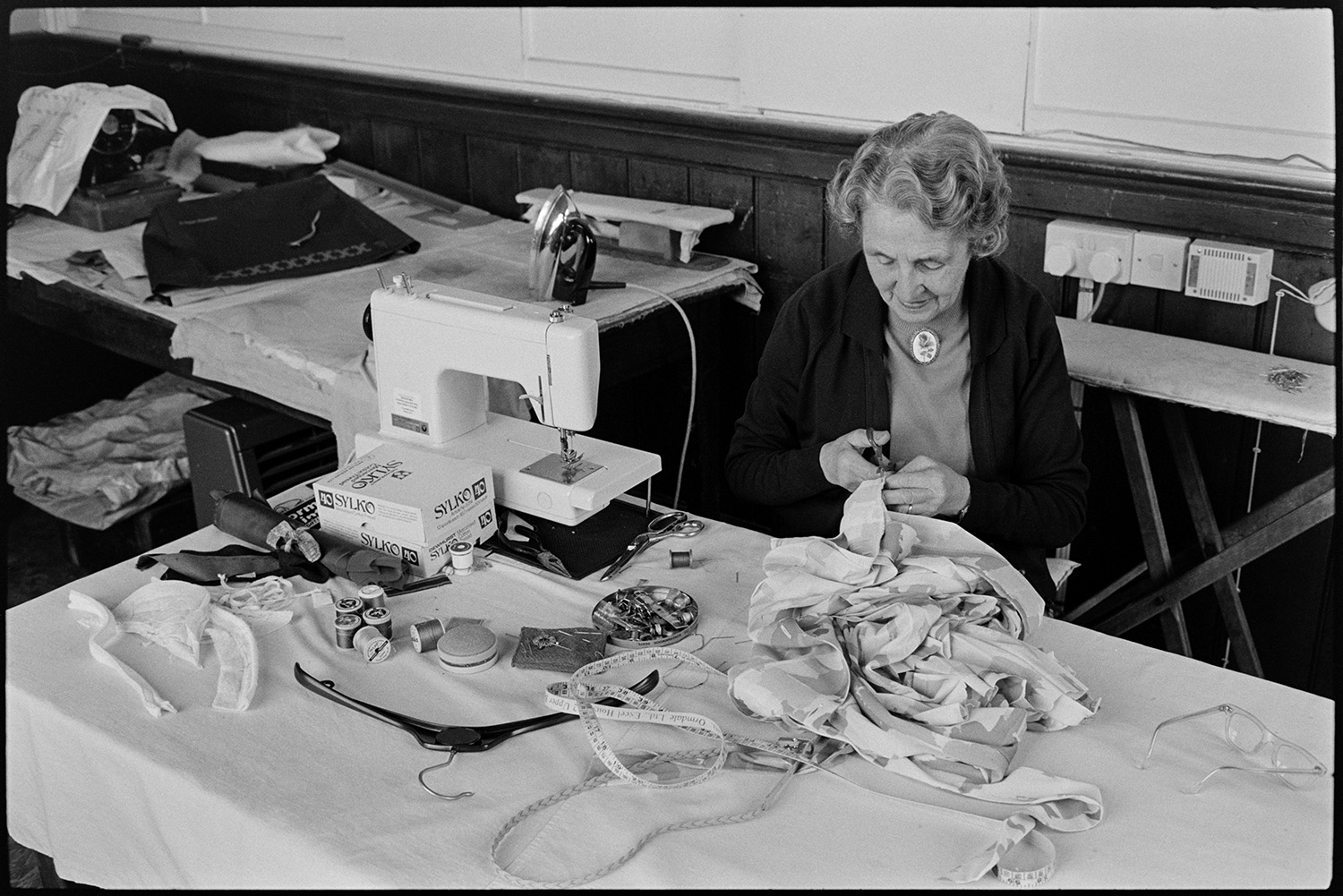 Woman sewing in workshop above clothes shop. 
[A woman sewing items of clothing using a sewing machine in a workshop above Trapnells clothes shop in Bideford High Street. A tape measure and reels of cotton are on the desk in front of her.]
