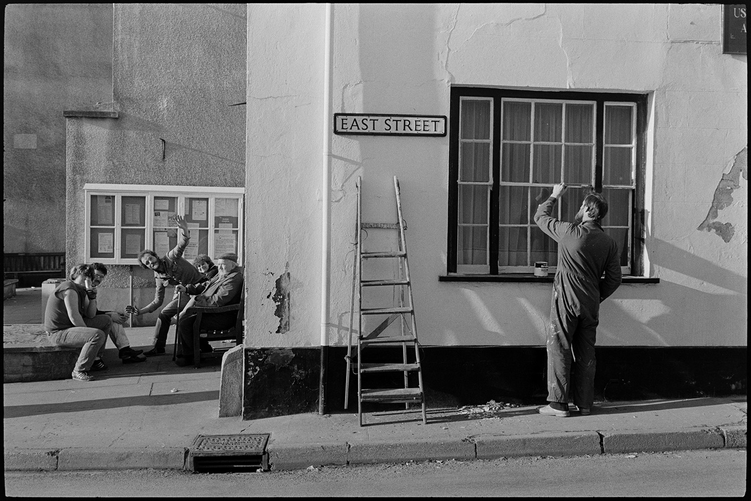 Street scenes with painter working on window, cyclists and passers by. Public house. 
[A man painting the window frames of the Red Lion Hotel  in East Street, Chulmleigh. His ladder is lent against the wall next to him. People sat on a bench, by a noticeboard next to the pub, are waving at the camera.]