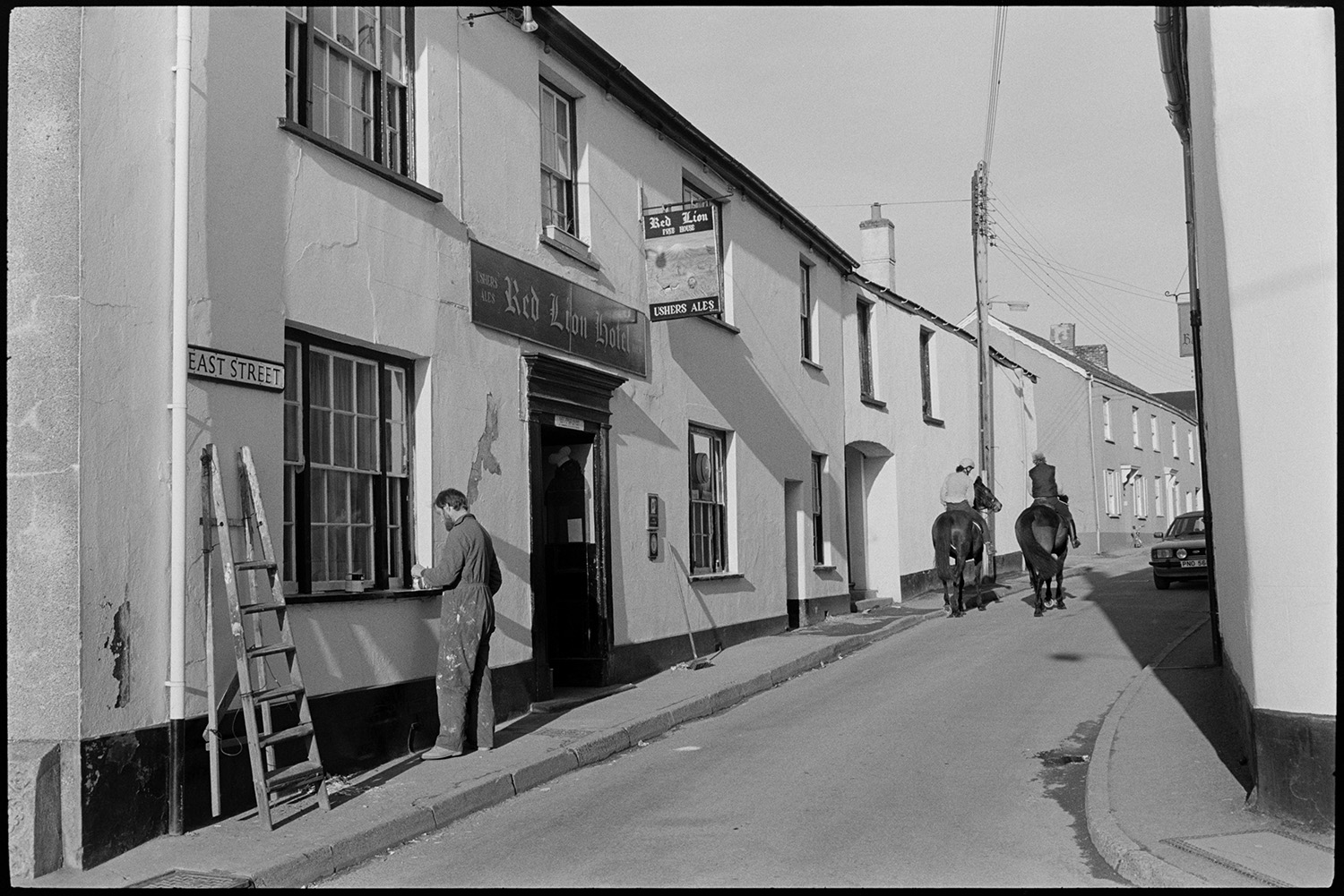 Street scenes with painter working on window, cyclists and passers by. Public house. 
[A man painting the window frames of the Red Lion Hotel  in East Street, Chulmleigh. His ladder is lent against the wall next to him. Two mounted horse riders can be seen further up the street.]