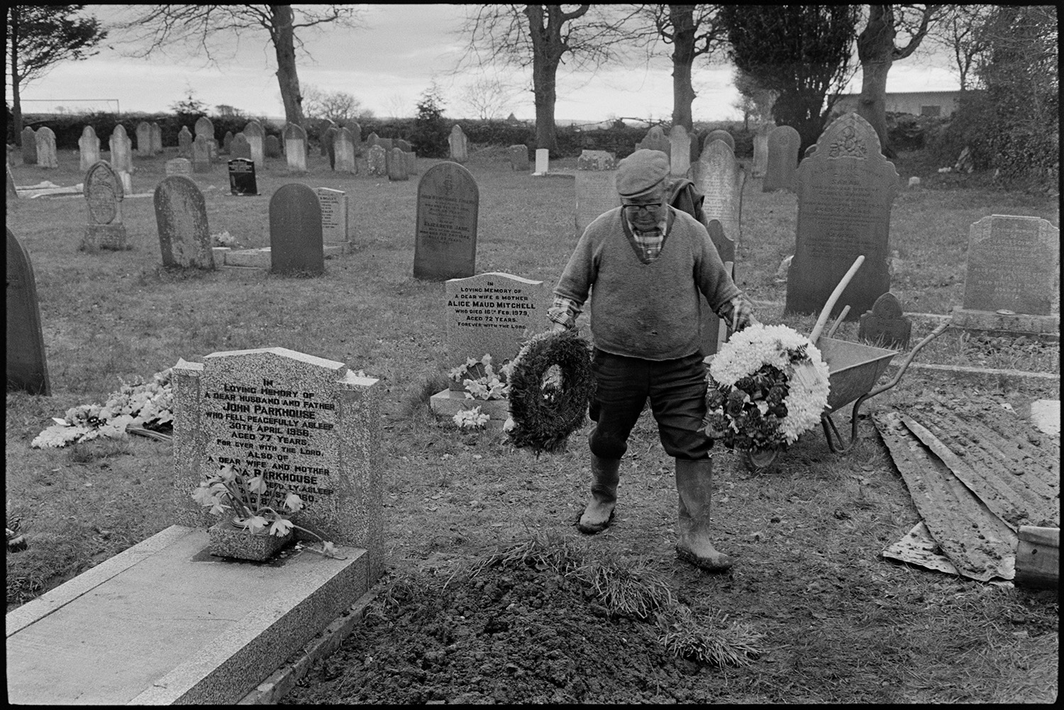 Gravedigger arranging flowers after funeral. 
[Reginald Rice arranging flowers on Archie Parkhouse's grave, after filling in the grave with earth, in Dolton churchyard. His wheelbarrow can be seen behind him. Other gravestones are visible, including Archie's parents' grave which is next to his.]