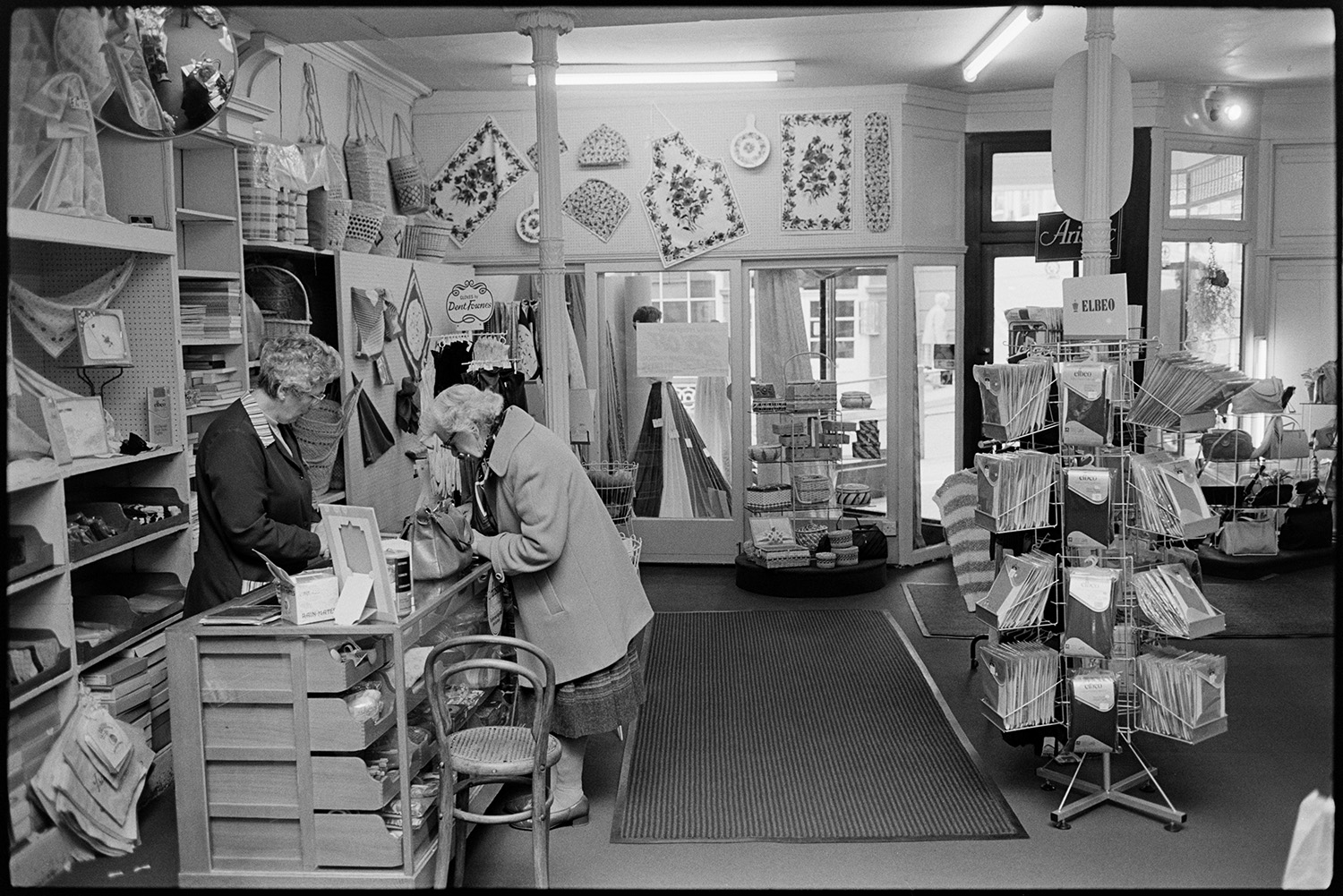 Interior of clothes shop customers being served at various counters. 
[A woman buying good at a counter in Trapnells clothes shop in Bideford High Street. Various items are displayed around the counter, including tea towels, aprons and bags.]