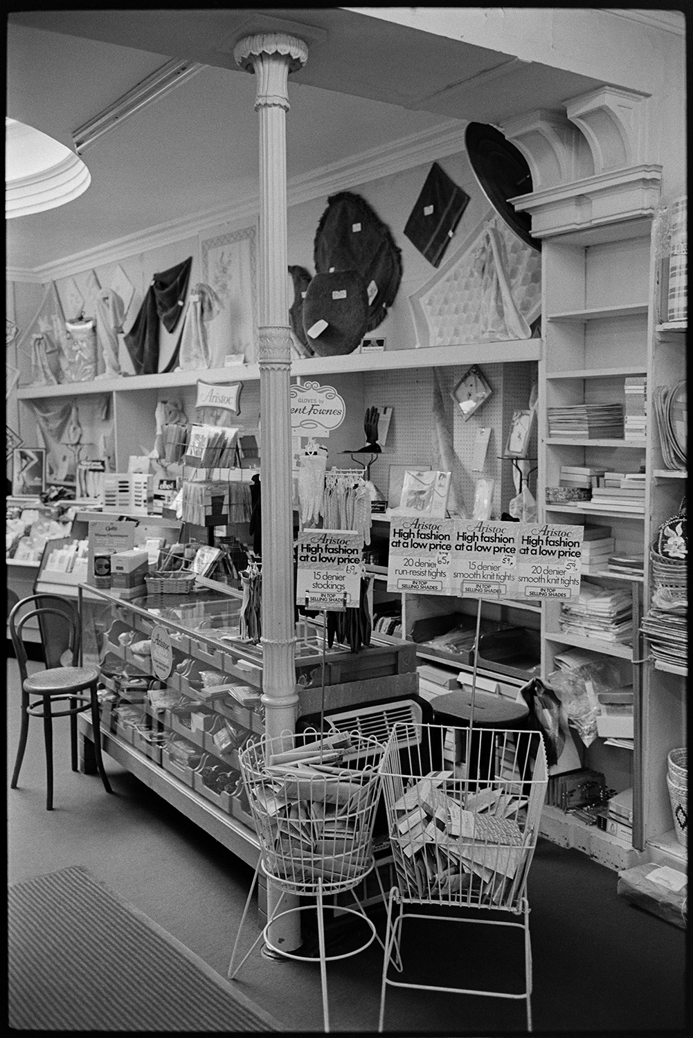 Interior of clothes shop customers being served at various counters. 
[A shop counter in Trapnells clothes shop in Bideford High Street. Various items are on display including tights and throws.]