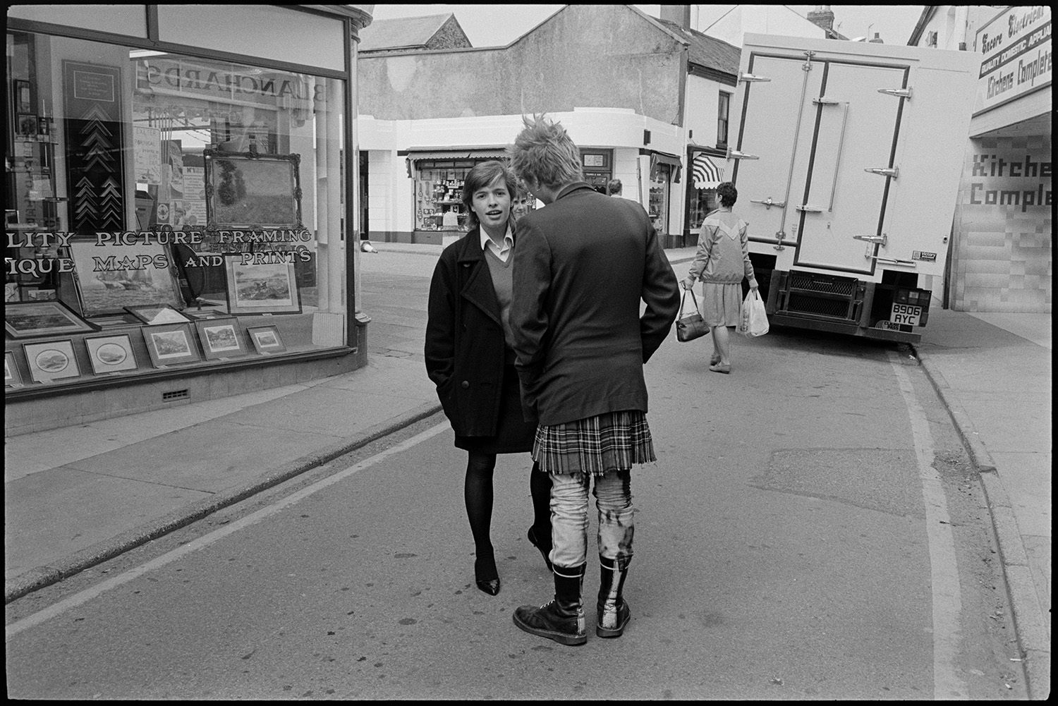 A young woman talking to a punk in a street outside a picture framing shop, possibly in Bideford. Other shop fronts and a lorry can be seen in the background.