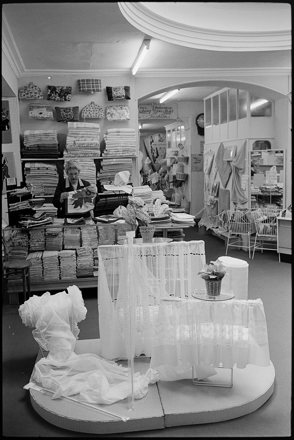Interior of clothes shop. Various counters, proprietor chatting, window and hat display. 
[A woman folding a tea towel behind a counter in Trapnells clothes shop in Bideford High Street. Stacks of towels and bags are on display behind her. In the foreground is a display of net curtains.]