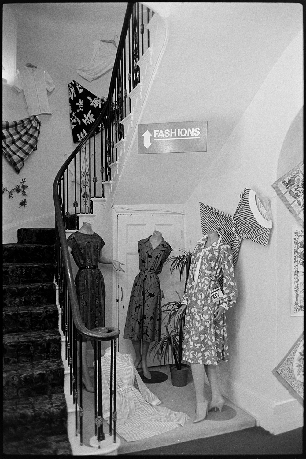 Interior of clothes shop. Various counters, proprietor chatting, window and hat display. 
[A display of women's dresses by a staircase in Trapnells clothes shop in Bideford High Street. The dresses are on mannequins. Skirts and blouses are displayed on the wall by the stairs.]