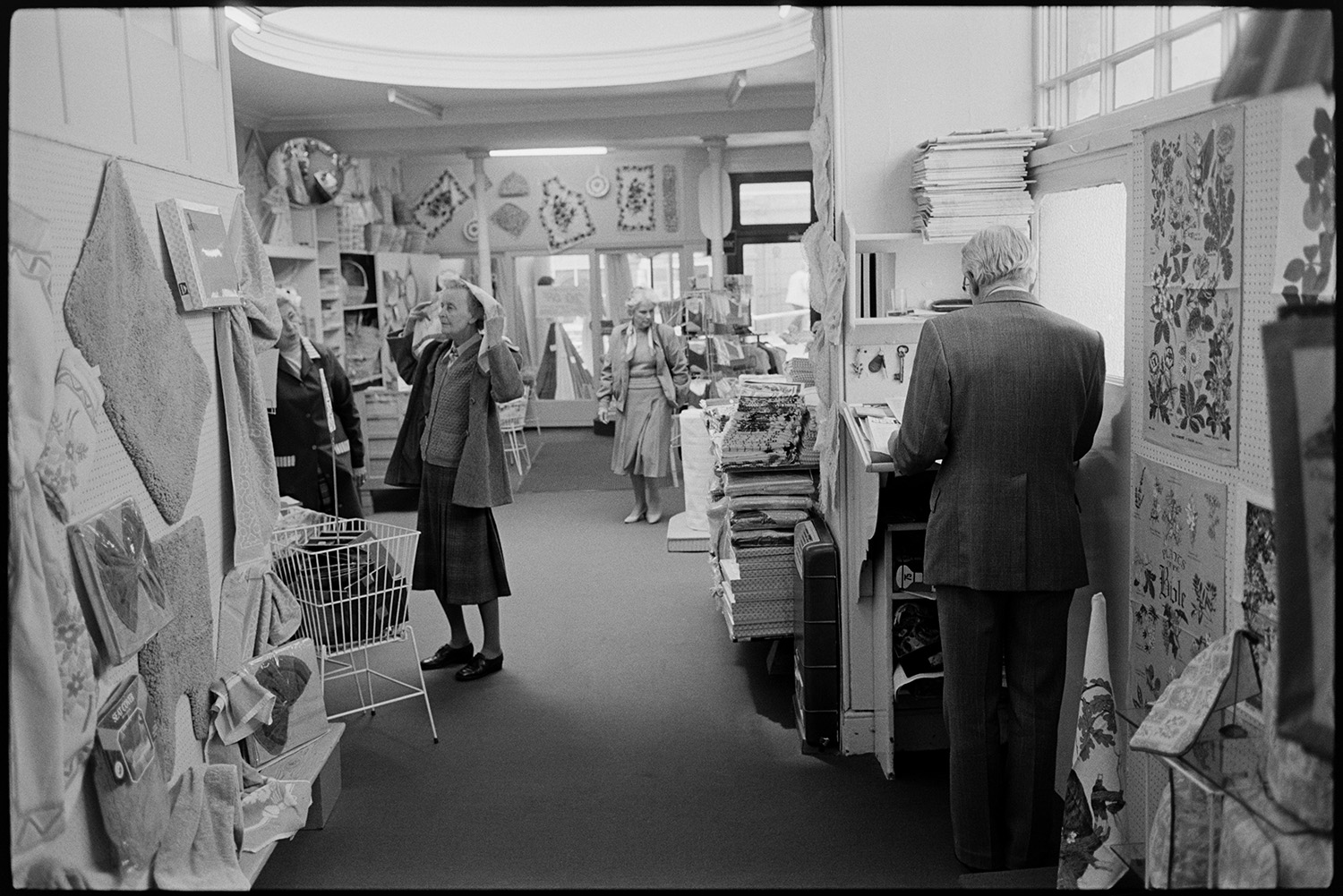 Interior of clothes shop. Various counters, proprietor chatting, window and hat display. 
[Women shopping in Trapnells clothes shop in Bideford High Street. One woman is trying on a headscarf. A man is stood at a small desk in the foreground. Various goods are on display in the shop, including tea towels and bath mats.]