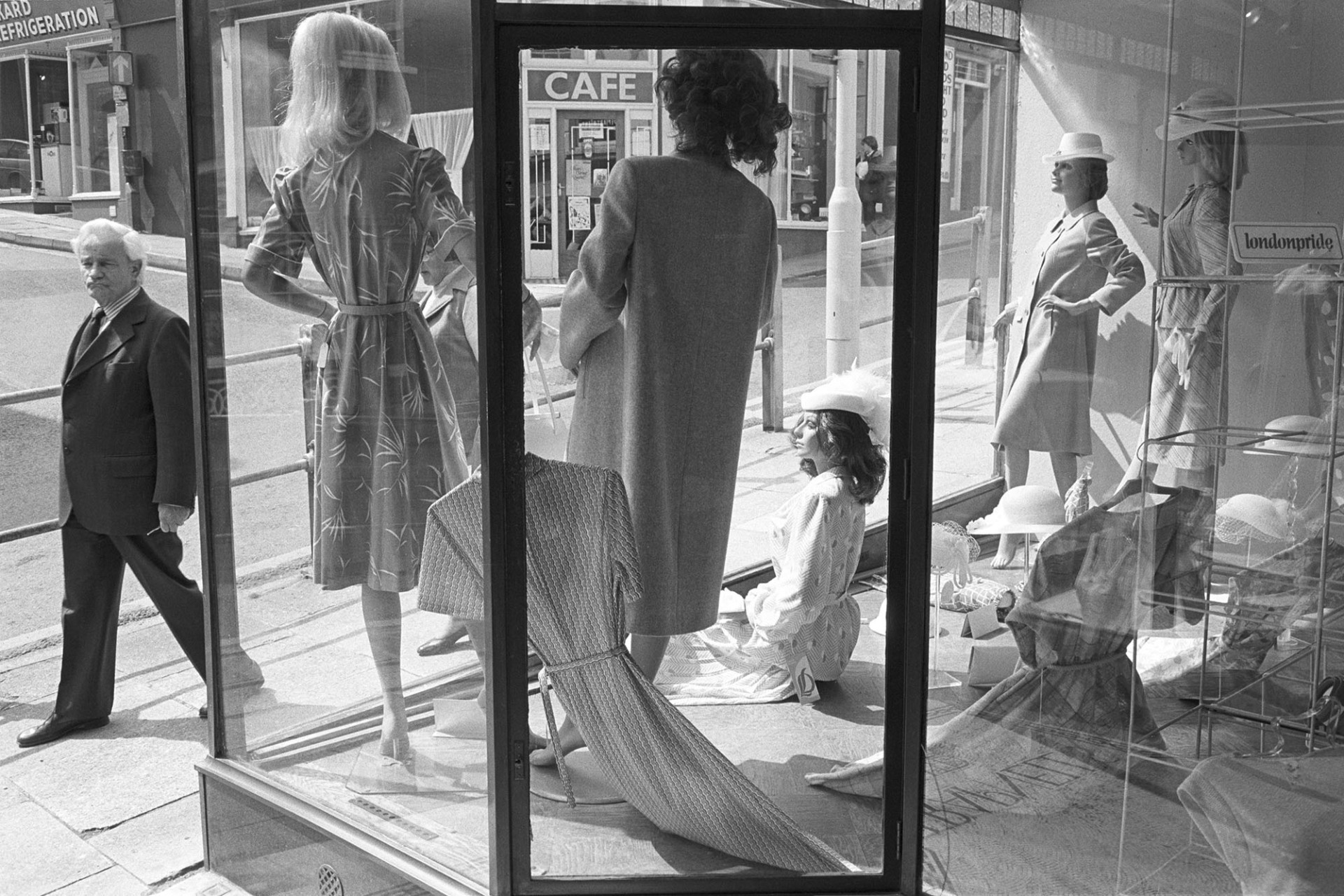 Window display of clothes shop with passer by. 
[A man walking past the shop window of Trapnells shop in Bideford High Street. The shop window is displaying women's clothes and hats on mannequins.]