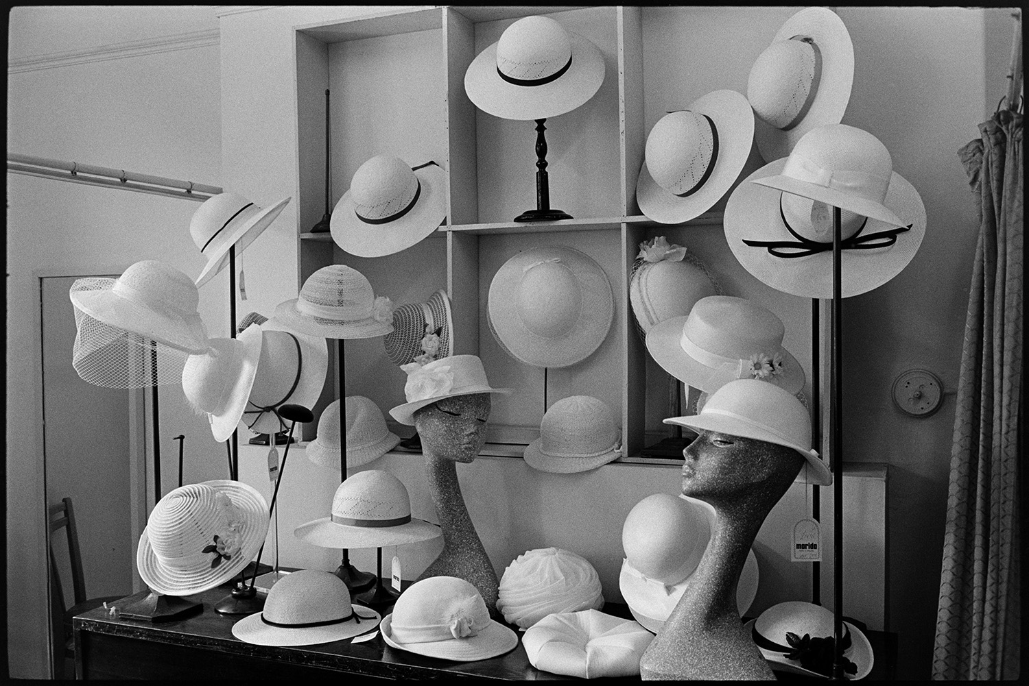Interior of clothes shop. Various counters, proprietor chatting, window and hat display. 
[A display of hats in Trapnells clothes shop in Bideford High Street. Some of the hats are on models or mannequins.]