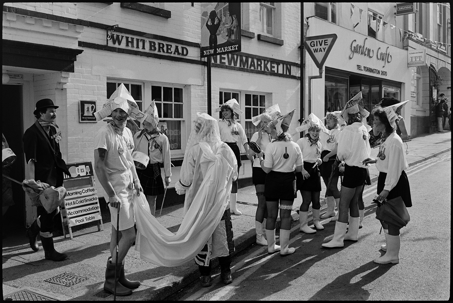 May Fair floats. Fairground procession. Generator lorry. Merry-go-round. 
[A group of men and women in fancy dress wearing paper hats and wellington boots at Torrington May Fair. They are stood in a street outside the Newmarket Inn and Garden Craft shop.]