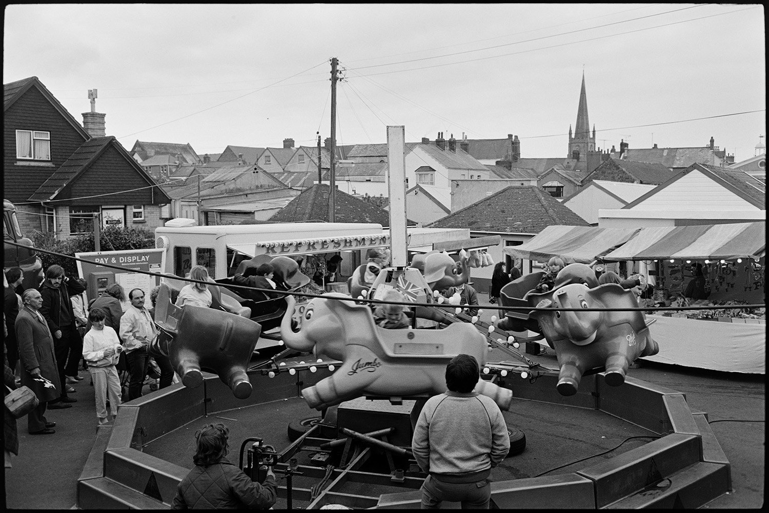 May Fair floats. Fairground procession. Generator lorry. Merry-go-round. 
[A children's merry-go-round 'Dumbo' ride at Torrington May Fair. Parents are watching children on the ride. Other stalls at the fairground, including a refreshment van, can be seen in the background.]