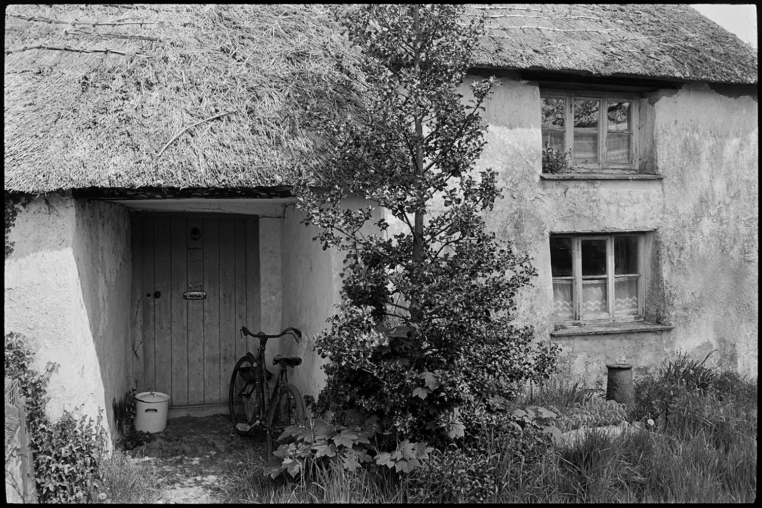 Street scenes, house with porch, thatched farmhouse and door, people chatting, church tower. 
[A bicycle parked in the porch outside the front door to a thatched farmhouse in Northlew.]