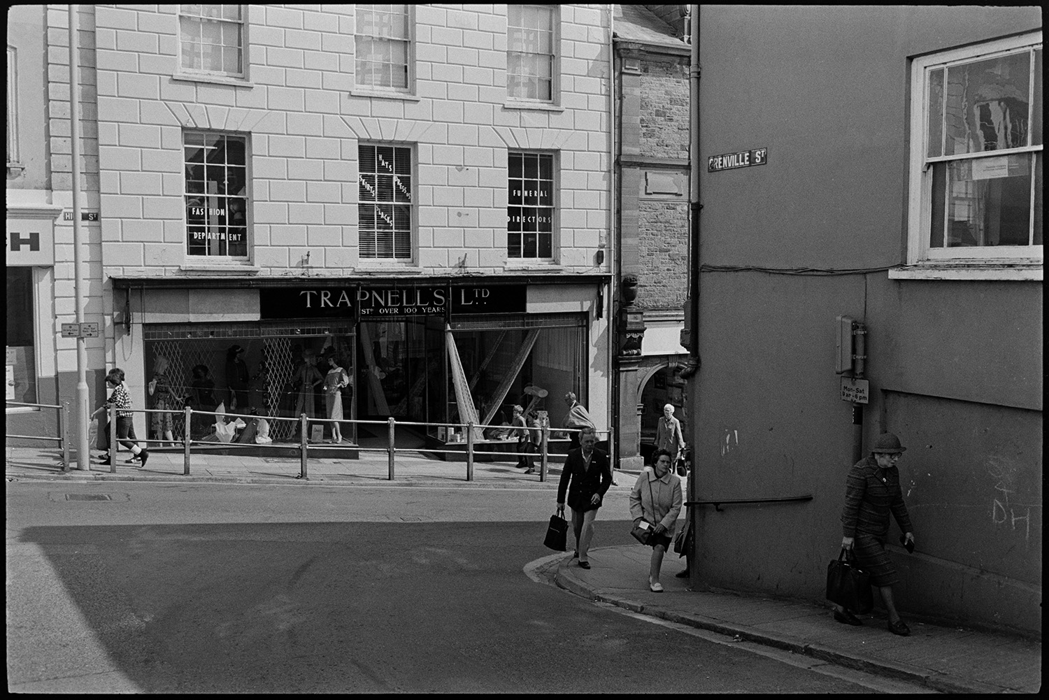 Front of former clothes shop with  sign. 
[A view from Grenville Street onto the High Street in Bideford, showing the shop front of Trapnells clothes shop. Mannequins can be seen in the shop window. Shoppers and passers by are walking along the streets.]