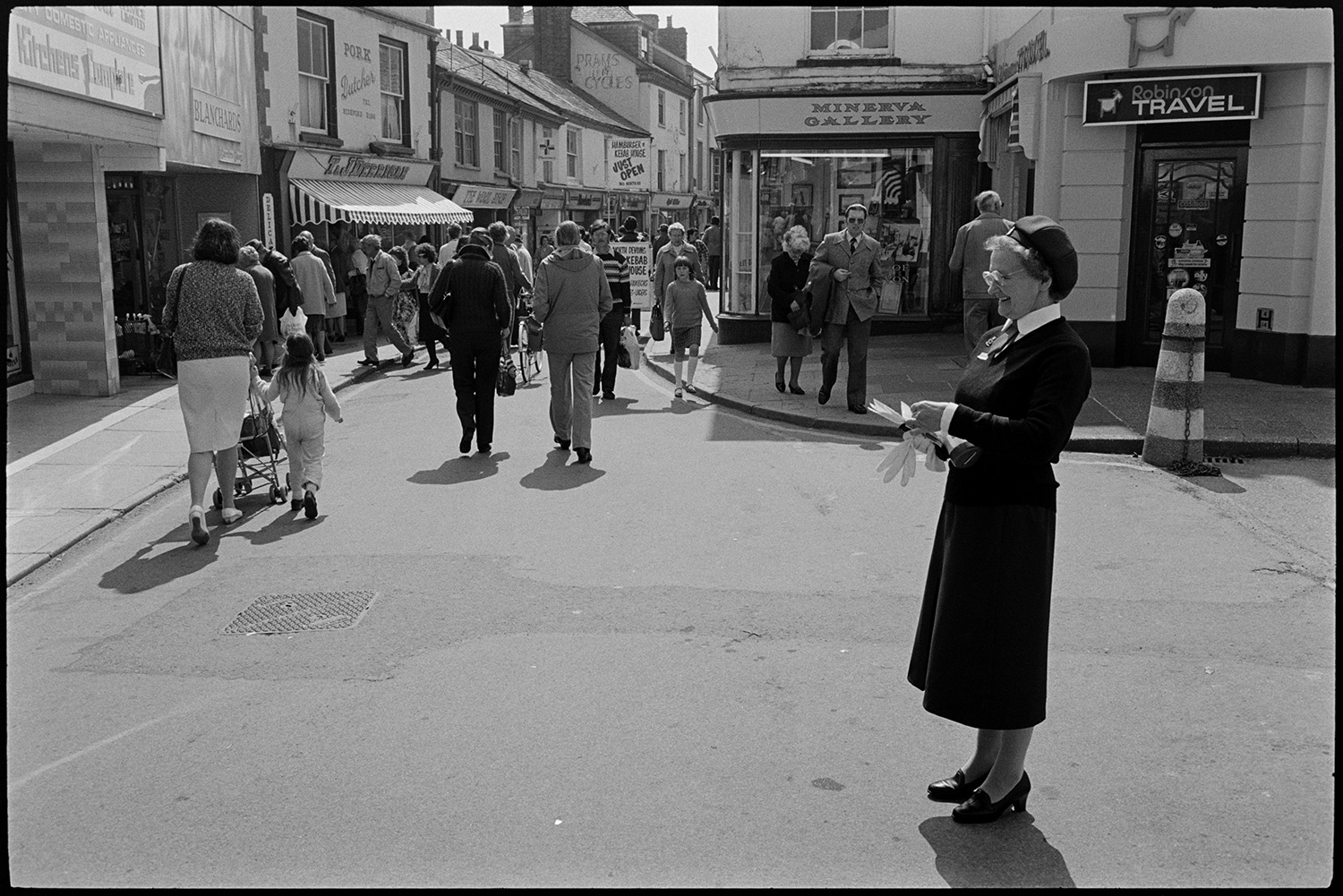 Street scenes. 
[A woman stood at a road junction in Mill Street, Bideford. Shoppers can be seen further along the street with various shop fronts, including Blanchard's, Robinson Travel and Minerva Gallery.]