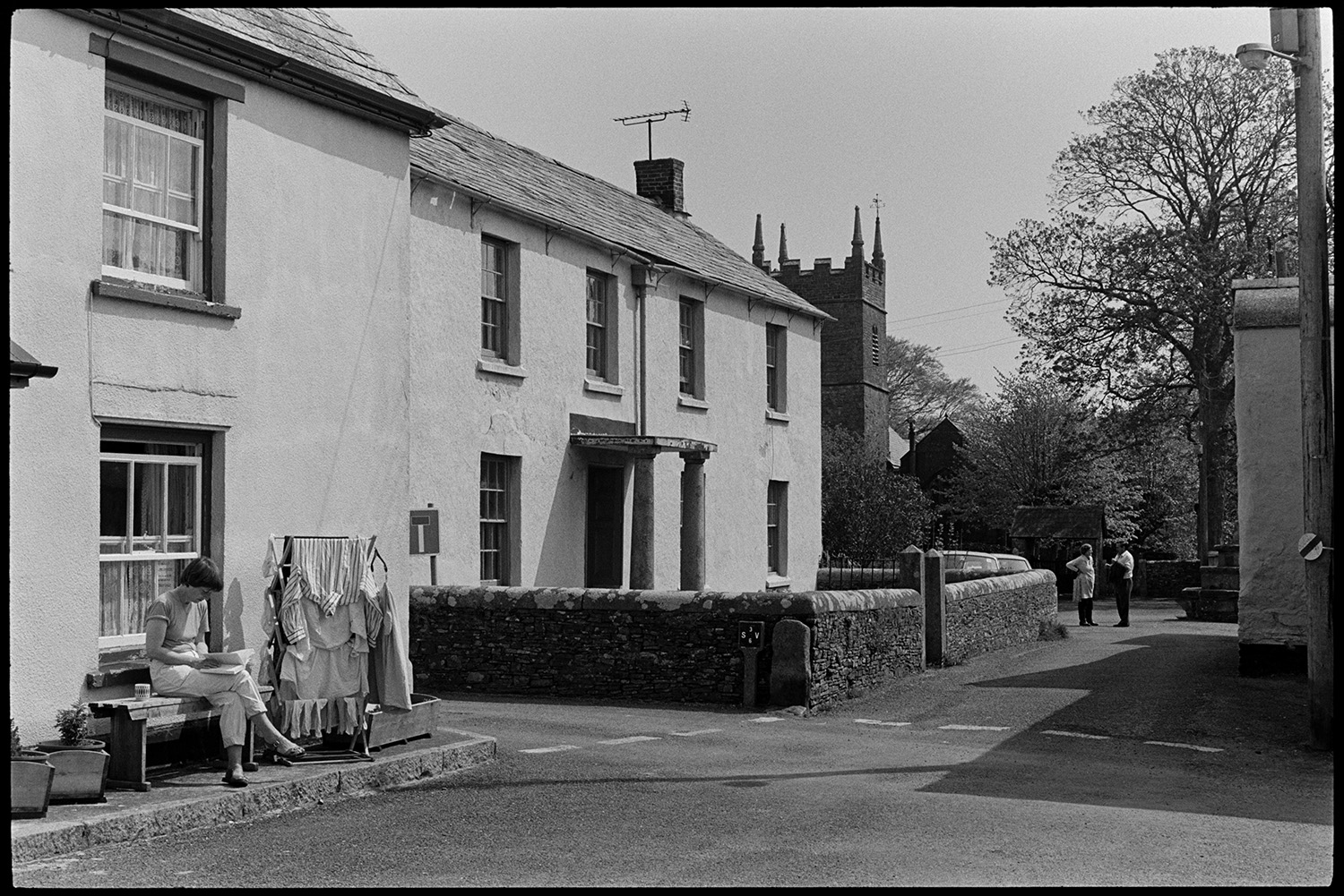 Street scenes, house with porch, thatched farmhouse and door, people chatting, church tower. 
[A woman sat on a bench next to a clothes airer drying washing, outside a house in Northlew. A house with a porch held up by pillars and the church tower can be seen in the background, along with two people talking in the street.]